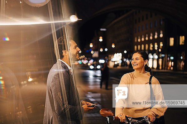 Lächelnde junge Frau mit Fahrrad schaut einen männlichen Freund an  während sie nachts in der Stadt steht