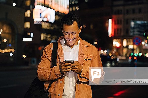 Smiling young man texting through phone while standing in illuminated city at night