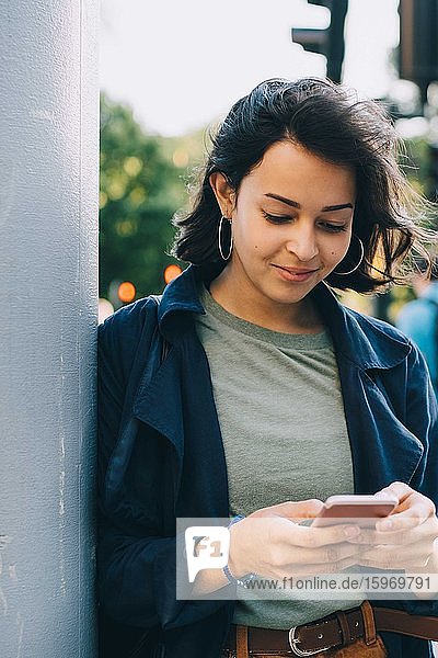 Lächelnde junge weibliche Textnachrichten  während sie an einer Säule in der Stadt stehen