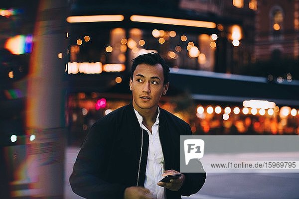 Young man with phone looking away while standing in illuminated city at night