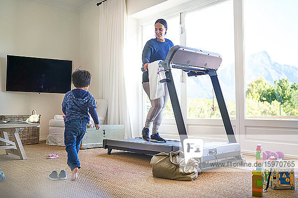 Junge spielt im Wohnzimmer  während die Mutter auf dem Laufband trainiert