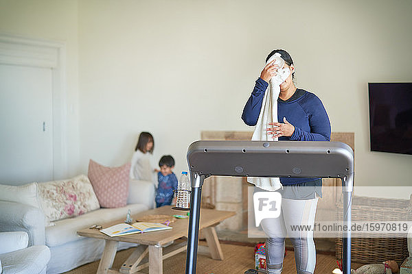Mutter auf Laufband wischt im Wohnzimmer mit Kindern den Schweiss vom Gesicht