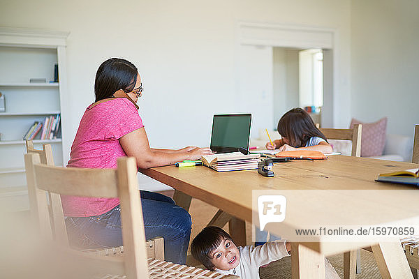 Mutter arbeitet am Laptop mit spielenden Kindern und macht Hausaufgaben