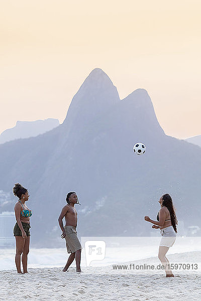 Ein junger schwarzer Brasilianer spielt mit zwei Freundinnen Fussball am Strand von Ipanema mit dem Dois Irmaos-Gebirge in der Ferne  Rio de Janeiro  Brasilien  Südamerika