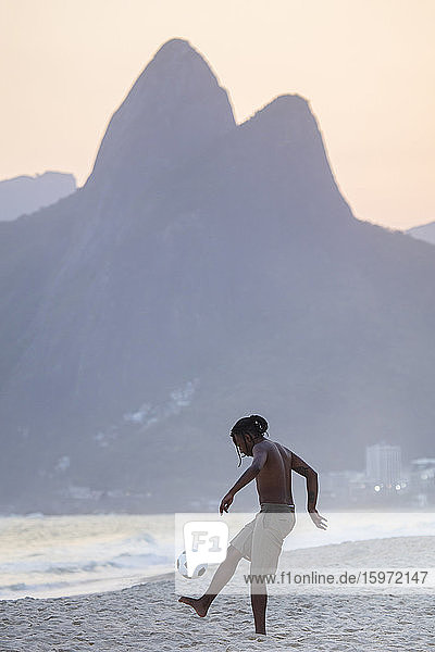 Ein junger schwarzer Brasilianer spielt Fussball am Strand von Ipanema mit dem Dois Irmaos-Gebirge in der Ferne  Rio de Janeiro  Brasilien  Südamerika