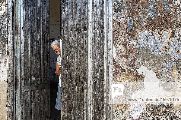 Frau am Eingang ihres Hauses mit einer verfallenen Fassade aus bröckelndem Putz und einer verwitterten Holztür  Gibara  Kuba  Mittelamerika