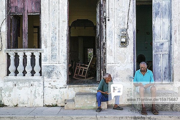 Zwei Männer unterhalten sich an den Türschwellen ihrer Häuser mit verfallenen Fassaden aus bröckelndem Putz und verwitterten Holztüren  Gibara  Kuba  Mittelamerika