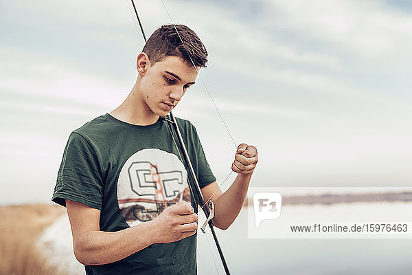 Jugendlicher bindet Haken an Angelrute  während er am Seeufer vor bewölktem Himmel steht