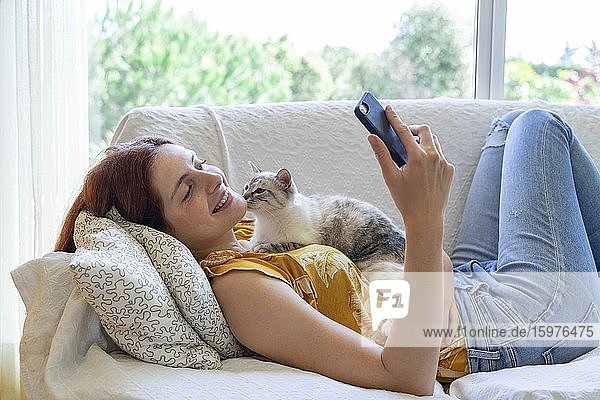 Glückliche junge Frau  die mit ihrer Katze zu Hause auf der Couch liegt und ihr Smartphone benutzt