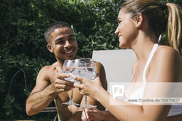 Lächelnder junger Mann stößt mit seiner Freundin auf ein Getränk an  während er auf einem Liegestuhl sitzt