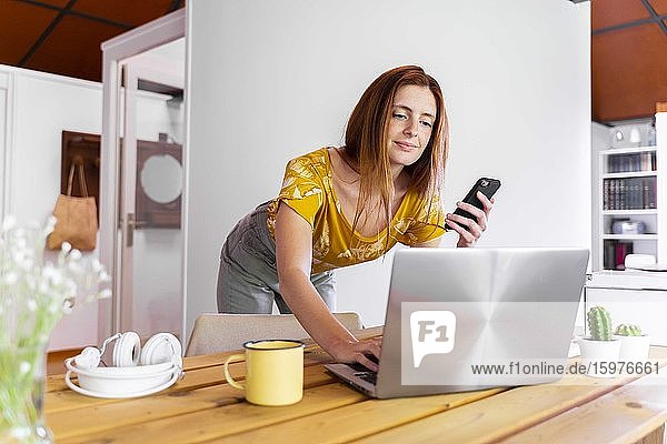 Frau hält Smartphone und arbeitet über Laptop auf dem Schreibtisch zu Hause während der Ausgangssperre