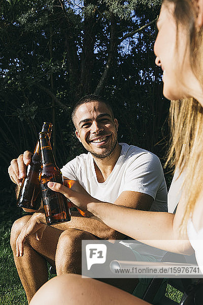 Lächelnder Mann stößt mit seiner Freundin auf eine Bierflasche an  während er in einem Touristenort sitzt