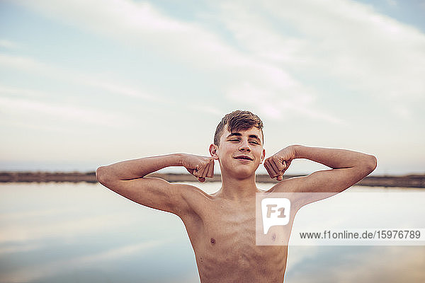 Lächelnder  hemdsärmeliger Teenager  der Muskeln anspannt und wegschaut  während er mit einem See und einem bewölkten Himmel im Hintergrund steht