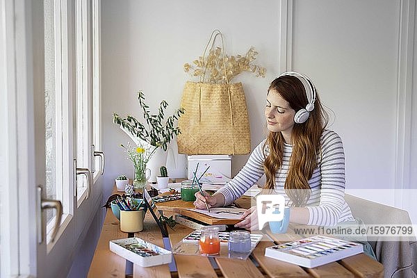 Young woman listening music though headphones while painting on table at home