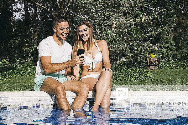 Smiling man showing smart phone to girlfriend while sitting against trees at poolside