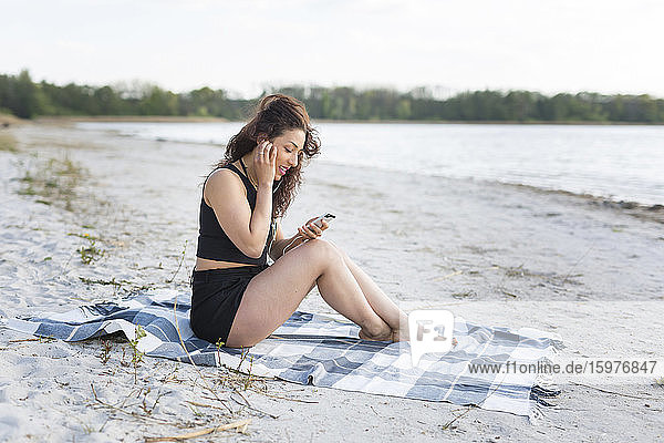 Young woman sitting on blanket on the beach using earphones and smartphone