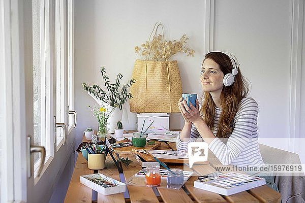 Nachdenkliche junge Frau hält Kaffeetasse und hört Musik  während sie zu Hause auf dem Tisch malt