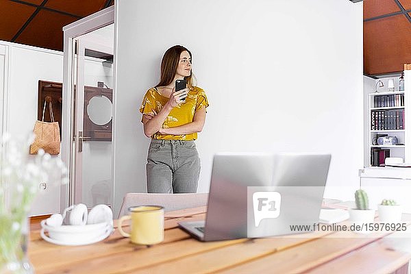Nachdenkliche Frau mit Smartphone an der Wand stehend zu Hause