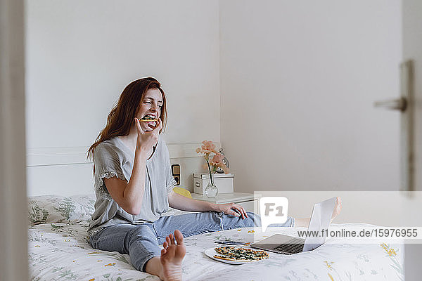 Young woman eating pizza while watching movie on laptop in bedroom at home