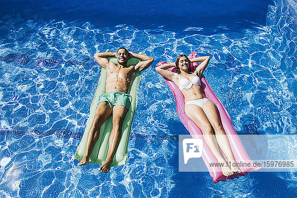 Lächelndes junges Paar mit Händen hinter dem Kopf  das sich auf Luftmatratzen im Schwimmbad entspannt