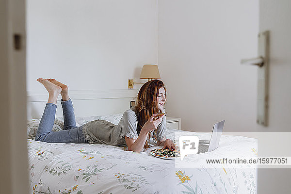 Frau isst Pizza  während sie im Schlafzimmer einen Film auf dem Laptop schaut