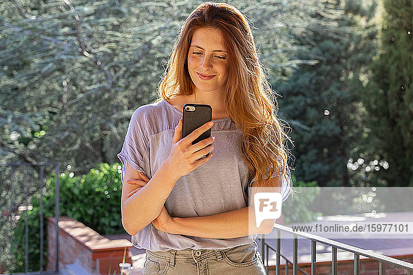 Lächelnde rothaarige Frau überprüft ihr Smartphone auf der Terrasse