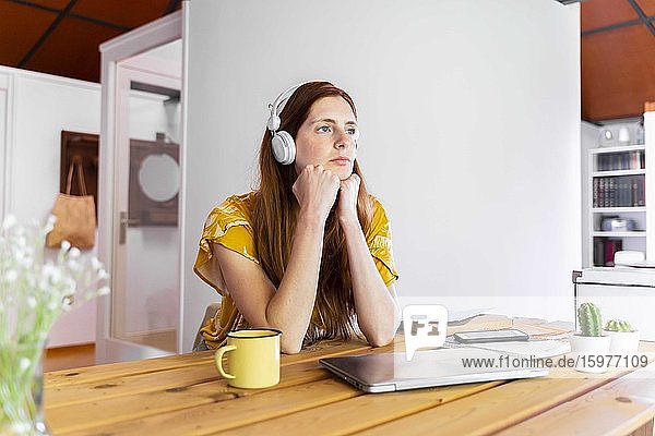 Thoughtful young woman with hands on chin sitting at desk against wall