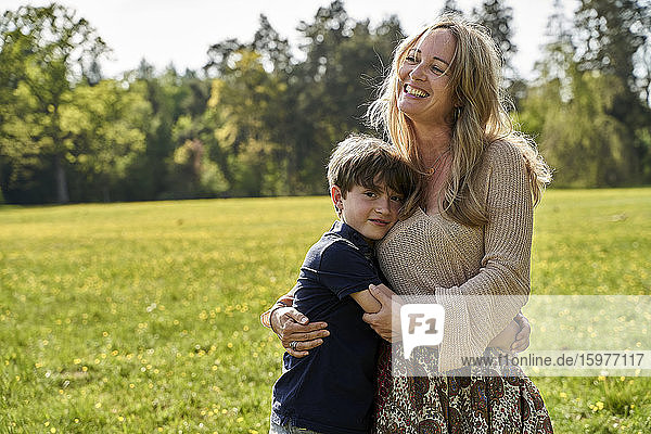 Junge umarmt lächelnd blonde Mutter  während auf Gras während sonnigen Tag stehen