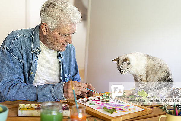 Älterer Mann malt auf Papier,  während die Katze zu Hause auf dem Tisch sitzt