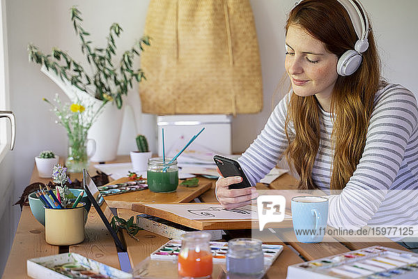 Junge Frau hört Musik über ihr Smartphone  während sie zu Hause am Tisch malt