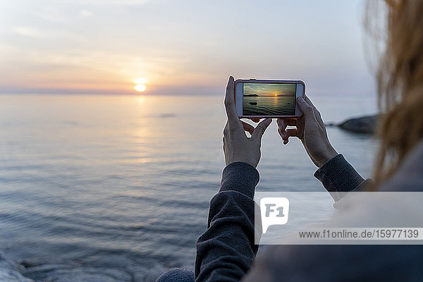 Spanien  Costa Brava  Nahaufnahme einer Frau  die mit ihrem Smartphone das Meer bei Sonnenaufgang fotografiert