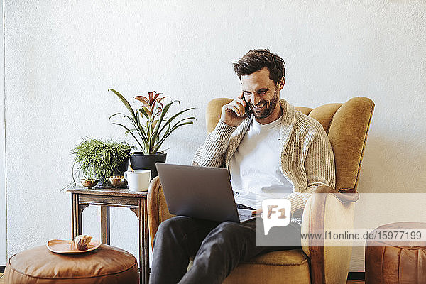 Lächelnder Mann am Telefon  der zu Hause auf einem Sessel sitzt und auf einen Laptop schaut
