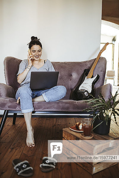 Porträt einer lächelnden jungen Frau am Telefon  die zu Hause auf der Couch sitzt und auf den Laptop schaut