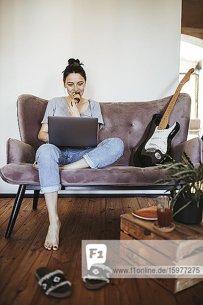 Junge Frau sitzt zu Hause auf der Couch und isst ein Croissant  während sie auf ihren Laptop schaut