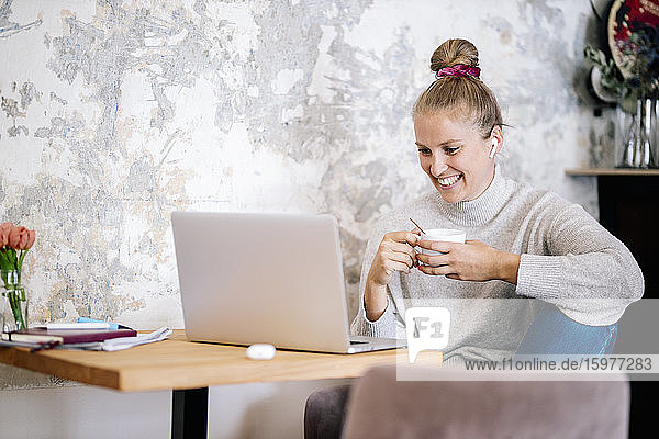 Smiling blond woman sitting on armchair at home having video chat with laptop and earphones