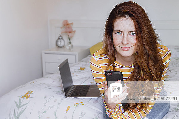 Schöne Frau mit grauen Augen und langen braunen Haaren sitzt mit Handy auf Schlafzimmer zu Hause