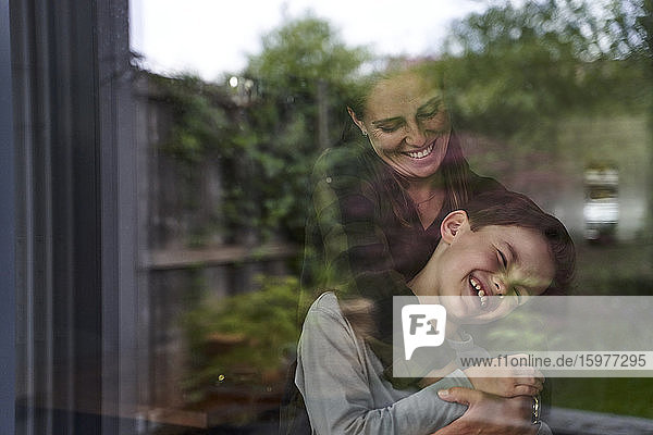 Frau umarmt fröhlichen Sohn zu Hause durch Fenster gesehen