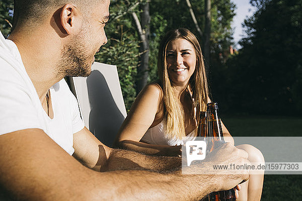 Lächelndes junges Paar stößt mit Bierflaschen an  während es in einem Touristenort sitzt