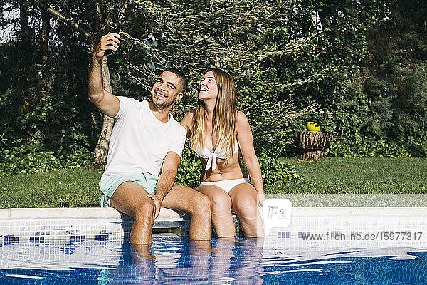 Lächelnder Mann  der ein Selfie mit seiner Freundin über ein Smartphone macht  während er am Pool gegen Bäume sitzt