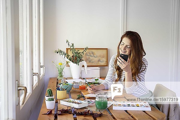Glückliche junge Frau malt  während sie ihr Smartphone auf dem Tisch zu Hause hält