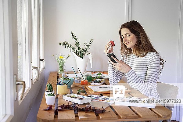Junge Frau isst eine Erdbeere und benutzt ihr Smartphone  während sie zu Hause auf dem Tisch malt