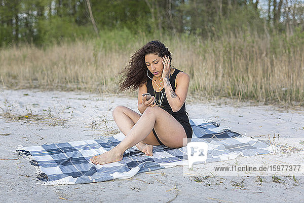 Junge Frau sitzt auf einer Decke am Strand und benutzt Kopfhörer und Smartphone