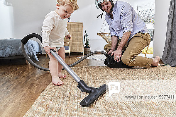 Mann schaut auf niedlichen Kleinkind Sohn Reinigung Teppich mit Staubsauger im Wohnzimmer zu Hause