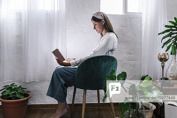 Entspannte Frau  die ein Buch liest  während sie auf einem Stuhl bei den Pflanzen zu Hause sitzt