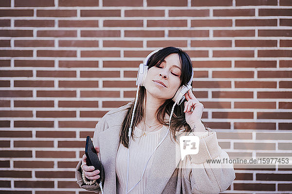 Porträt einer Frau mit geschlossenen Augen  die mit Kopfhörern und Smartphone vor einer Backsteinmauer Musik hört