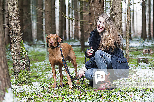 Lächelnde junge schöne Frau mit langen braunen Haaren hockt neben einem Hund an einem Baum im Wald