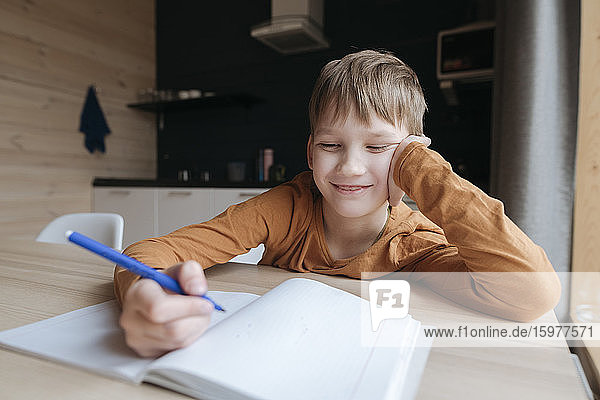 Smiling boy sitting at table writing into booklet at home