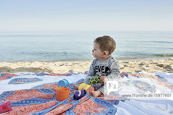 Cute boy with toys sitting on blanket at beach against sea