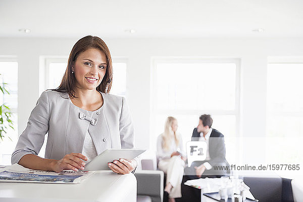 Glückliche Geschäftsfrau  die ein digitales Tablet hält  während Kollegen im Hintergrund im Büro diskutieren