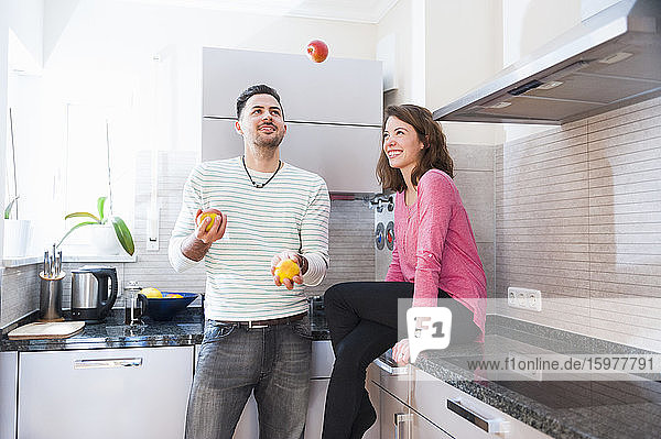 Happy woman looking at man juggling with fruits in kitchen